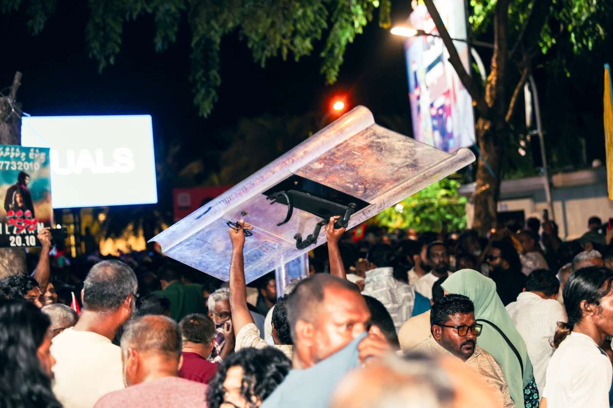 އޮކްޓޯބަރު 3، 2025: މުޒާހަރާގައި ބައިވެރިވި މީހަކު ފުލުހުން ޝީލްޑެއް އުފުލާލައިގެން ދަނީ. މިހާ ފަސޭހައިން ޝީލްޑެއް އަތުލެވުނު ނަމަވެސް، ފުލުހުންގެ ސަފުތައް ފޫ އަޅުވާލައިފައި މުޒާހަރާގެ ބައިވެރިންނަށް މަޖީދީ މަގުން ކުރިއަށް ނުދެވުނު. ސުލުހަވެރި މުޒާހާރާ އަށް ހުރަސް އެޅުމަށް ފަހު ދިފާއު އަކަށް ފުލުހުން ނެގީ، މަޖީދީ މަގުން ހިނގަން ހުއްދަ ނުދީ އޮތް އޮތުން. ނަމަވެސް ސުލުހަވެރި މުޒާހަރާއެއް ނަމަ ފުލުހުން ހުރަސް ނާޅާނެ ކަމަށް ފުލުހުން ބުނެފައިވާ ކަމަށް އެމްޑީޕީން ބުނޭ. -- ފޮޓޯ: މިރާޝް ނާޝިމް