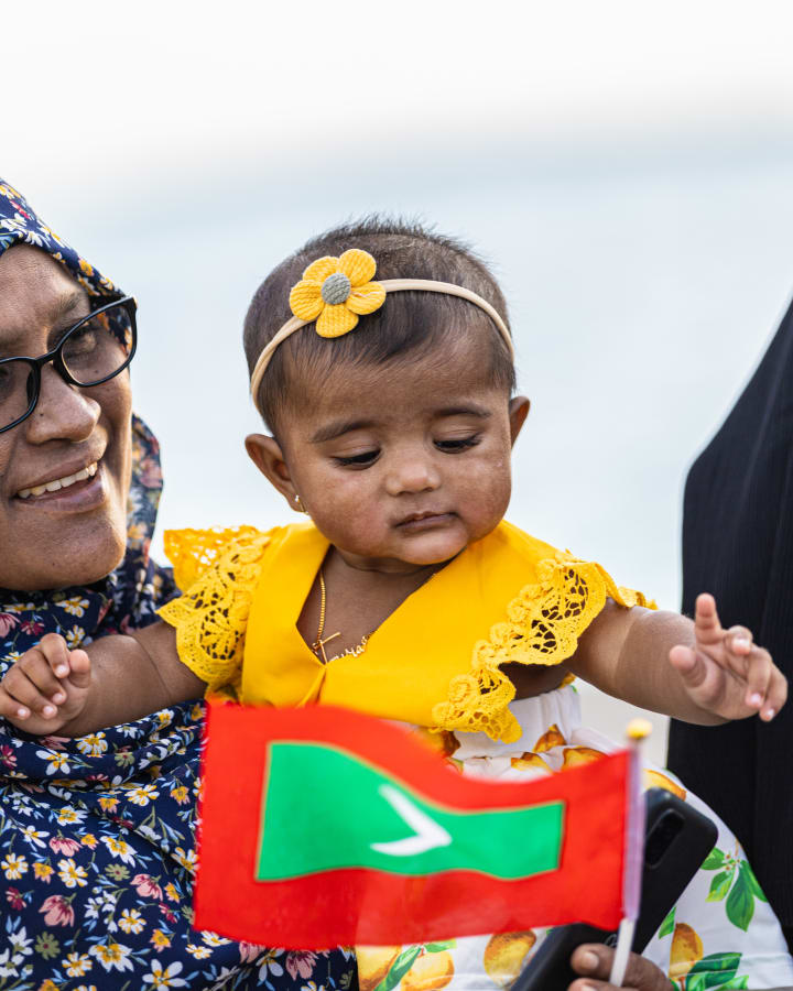 އޯގަސްޓު 7، 2023: ދ. ހުޅުދެލީގައި ރައީސް ސޯލިހަށް މަރުހަބާ ކިޔަން ނުކުމެ ހުރި އަންހެނަކު އުރައިގެން ހުރި ކުޑަ ކުއްޖާ ގައުމީ ދިދައާ ގުޅެނީ -- ފޮޓޯ: އަބްދުﷲ އިޔާން / އަދަދު 