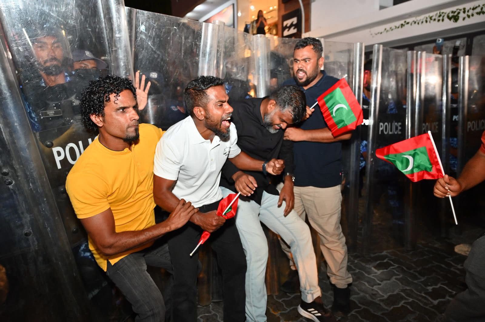 އޮކްޓޯބަރު 3، 2025: ރައީސް މުއިއްޒުގެ އިސްތިއުފާ އަށް ގޮވާލައި އެމްޑީޕީން ކުރި މުޒާހަރާ އަށް ފުލުހުން ހުރަސް އެޅުމުން ބައިވެރިން އެކަމާ ދެކޮޅު ހެދުމުގެ ގޮތުން ޝީލްޑުތައް ކައިރީގައި. މުޒާހަރާއިން ހައްޔަރުކުރި އަށް މީހުންގެ ތެރޭގައި ރައްޔިތުންގެ މަޖިލީހުގެ ކުރީގެ މެންބަރު ޔައުގޫބު (ރީނދޫ ޓީޝާޓުގައި) އަދި މާފުށީ ކައުންސިލްގެ ރައީސް ސޮލާހު (ކަޅު ޓީޝާޓުގައި ކޮޅަށް) ހައްޔަރުކުރި. -- ފޮޓޯ: އެމްޑީޕީ